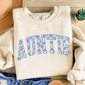 Peut inclure: Un sweat-shirt beige clair avec le mot "AUNTIE" imprim&eacute; en bleu floral sur la poitrine. Le sweat-shirt est pos&eacute; sur une surface en bois, avec un chapeau et un jean en arri&egrave;re-plan. Une petite plante succulente est &eacute;galement pr&eacute;sente.
