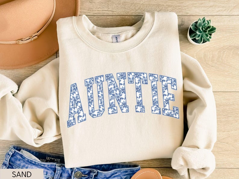 Peut inclure: Un sweat-shirt beige clair avec le mot "AUNTIE" imprim&eacute; en bleu floral sur la poitrine. Le sweat-shirt est pos&eacute; sur une surface en bois, avec un chapeau et un jean en arri&egrave;re-plan. Une petite plante succulente est &eacute;galement pr&eacute;sente.