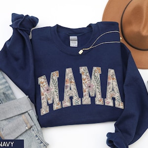 May include: Navy blue sweatshirt with the word "MAMA" in a floral design. A gold necklace and a brown hat are visible in the background. The sweatshirt is paired with denim jeans.