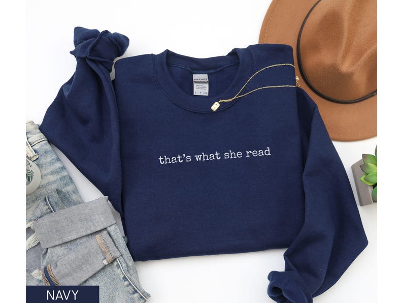 May include: Navy blue sweatshirt with the embroidered text "that's what she read." The long-sleeved top is paired with denim jeans and a gold necklace. A brown hat is visible in the background.