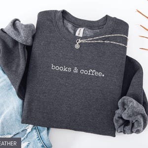 May include: Dark heather gray sweatshirt with the embroidered text "books & coffee." A silver necklace is draped across the front. The sweatshirt is paired with blue denim jeans. The image is well-lit and suitable for showcasing apparel.