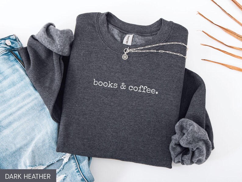 May include: Dark heather gray sweatshirt with the embroidered text "books & coffee." A silver necklace is draped across the front. The sweatshirt is paired with blue denim jeans. The image is well-lit and suitable for showcasing apparel.