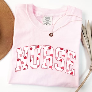 Pode incluir: Camiseta rosa com a palavra "NURSE" impressa em letras brancas com um padr&atilde;o de cora&ccedil;&otilde;es vermelhos. A camiseta est&aacute; dobrada e colocada sobre uma superf&iacute;cie branca.