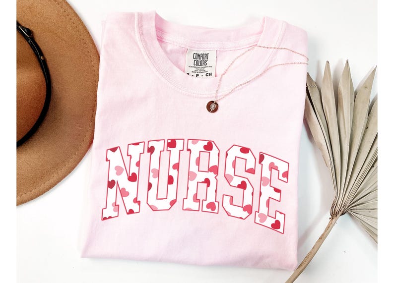 Pode incluir: Camiseta rosa com a palavra "NURSE" impressa em letras brancas com um padr&atilde;o de cora&ccedil;&otilde;es vermelhos. A camiseta est&aacute; dobrada e colocada sobre uma superf&iacute;cie branca.