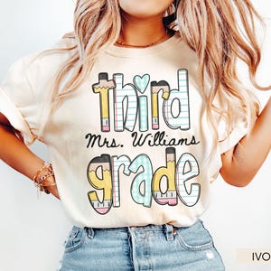 May include: Ivory-colored t-shirt with the words "third Mrs. Williams grade" in a playful, colorful font. The word "third" has a pencil design, and the word "grade" is also designed with pencils. The shirt is a teacher-themed item.