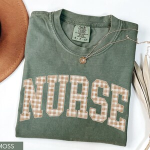 May include: A moss green t-shirt with the word "NURSE" in large, block letters with a tan and white gingham pattern. A gold necklace with a small pendant rests on the shirt. A brown hat is visible in the background.