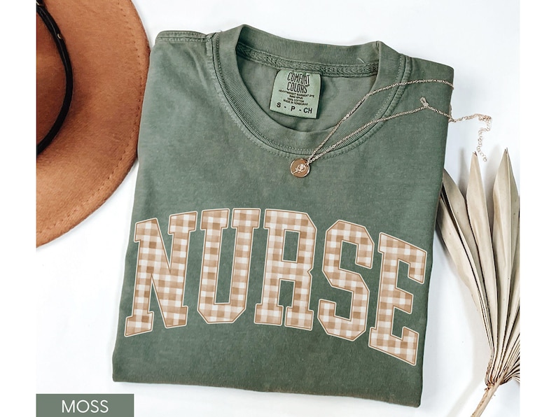 May include: A moss green t-shirt with the word "NURSE" in large, block letters with a tan and white gingham pattern. A gold necklace with a small pendant rests on the shirt. A brown hat is visible in the background.