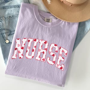 Pode incluir: Uma camiseta lavanda com a palavra "NURSE" impressa em branco com um padr&atilde;o de cora&ccedil;&otilde;es vermelhos e rosa.