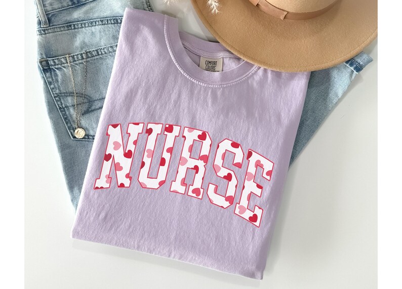 Pode incluir: Uma camiseta lavanda com a palavra "NURSE" impressa em branco com um padr&atilde;o de cora&ccedil;&otilde;es vermelhos e rosa.