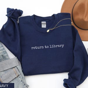 May include: Navy blue sweatshirt with the phrase "return to library" embroidered in white. The sweatshirt is paired with denim jeans, a gold necklace, and a brown hat. A small succulent plant is also visible.