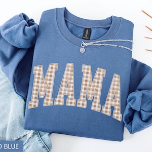 May include: Indigo blue sweatshirt with the word "MAMA" in a tan and white plaid pattern. A silver necklace with a small pendant is draped across the top. The sweatshirt is folded on top of a pair of blue jeans.