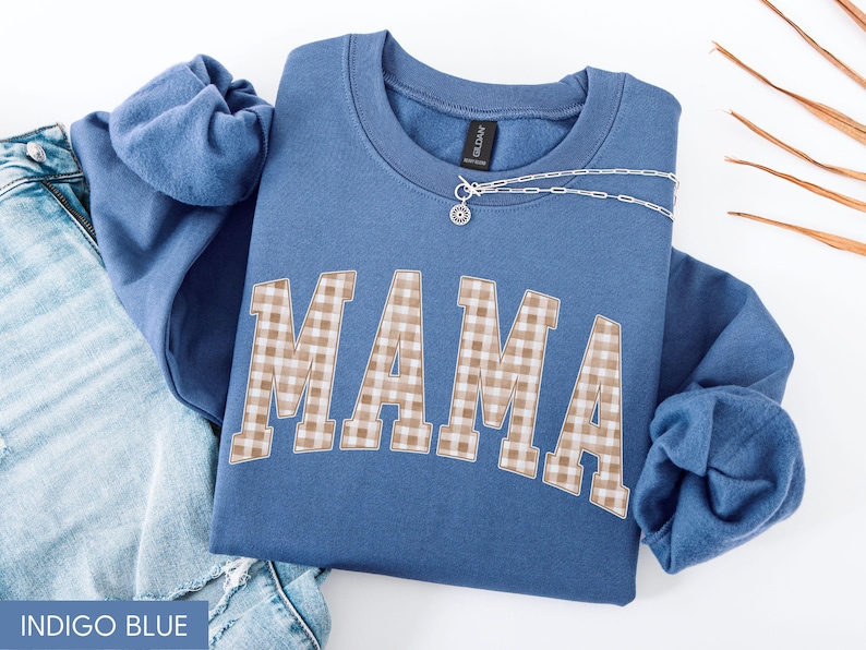 May include: Indigo blue sweatshirt with the word "MAMA" in a tan and white plaid pattern. A silver necklace with a small pendant is draped across the top. The sweatshirt is folded on top of a pair of blue jeans.