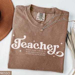 May include: Espresso-colored t-shirt with the word "Teacher" in a retro font, accompanied by the text "Some days I rock it, some days it rocks me, either way we're rockin". A silver necklace with a pendant is on the shirt.