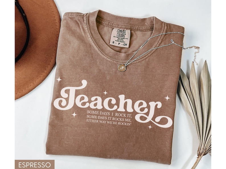 May include: Espresso-colored t-shirt with the word "Teacher" in a retro font, accompanied by the text "Some days I rock it, some days it rocks me, either way we're rockin". A silver necklace with a pendant is on the shirt.