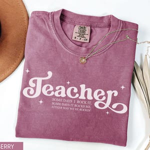 May include: Berry-colored t-shirt with the word "Teacher" in a white, retro font. The shirt also features the text "SOME DAYS I ROCK IT, SOME DAYS IT ROCKS ME, EITHER WAY WE'RE ROCKIN". A gold necklace and a brown hat are also visible.