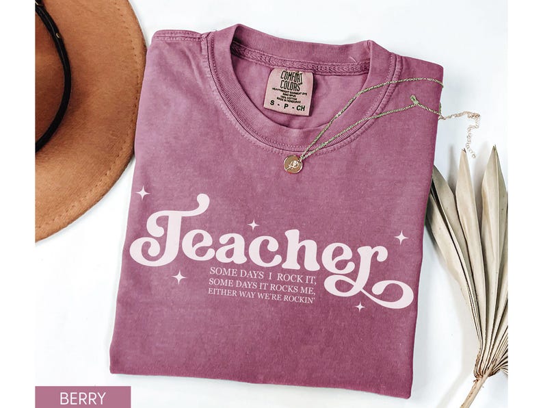 May include: Berry-colored t-shirt with the word "Teacher" in a white, retro font. The shirt also features the text "SOME DAYS I ROCK IT, SOME DAYS IT ROCKS ME, EITHER WAY WE'RE ROCKIN". A gold necklace and a brown hat are also visible.