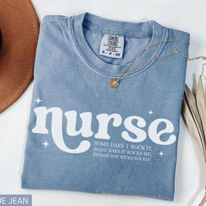 Pode incluir: Camiseta azul jeans com a palavra "nurse" em escrita branca. A camisa tem o texto "SOME DAYS I ROCK IT, SOME DAYS IT ROCKS ME, EITHER WAY WE'RE ROCKIN" abaixo da palavra "nurse". Um colar de ouro e um chap&eacute;u marrom tamb&eacute;m est&atilde;o na imagem.
