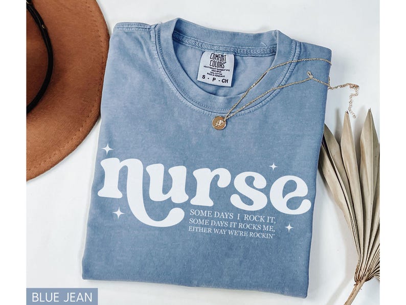 Pode incluir: Camiseta azul jeans com a palavra "nurse" em escrita branca. A camisa tem o texto "SOME DAYS I ROCK IT, SOME DAYS IT ROCKS ME, EITHER WAY WE'RE ROCKIN" abaixo da palavra "nurse". Um colar de ouro e um chap&eacute;u marrom tamb&eacute;m est&atilde;o na imagem.