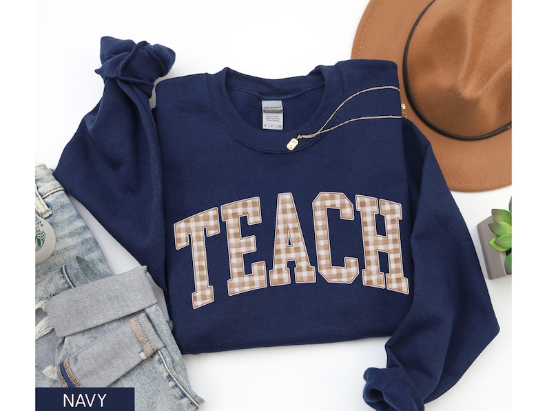 May include: Navy blue sweatshirt with the word "TEACH" in a tan and white plaid design. The sweatshirt is folded, with a pair of blue jeans and a brown hat in the background. A gold necklace is draped across the sweatshirt.