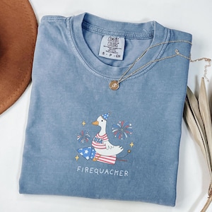 Op de afbeelding: Een lichtblauw T-shirt met een cartoonillustratie van een gans die een feestmuts draagt en op een vuurwerk zit. De gans houdt een vuurwerkstokje vast en er zijn vuurwerk in de achtergrond. De tekst "FIREQUACKER" is onder de gans afgedrukt.