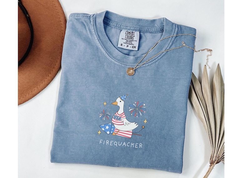 Op de afbeelding: Een lichtblauw T-shirt met een cartoonillustratie van een gans die een feestmuts draagt en op een vuurwerk zit. De gans houdt een vuurwerkstokje vast en er zijn vuurwerk in de achtergrond. De tekst "FIREQUACKER" is onder de gans afgedrukt.