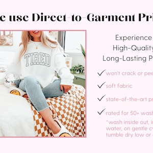 Pu&ograve; includere: Felpa bianca con la scritta "TIRED" in nero. L'immagine include anche il testo "We use Direct-to-Garment Print" e "Experience High-Quality, Long-Lasting Prints". L'immagine mostra una persona che indossa la felpa.