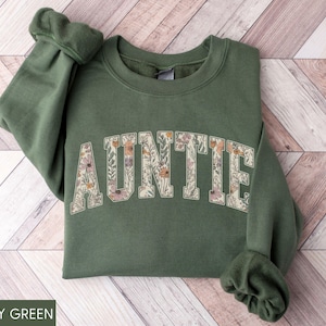 May include: Military green sweatshirt with the word "AUNTIE" in a floral design across the chest. The sweatshirt has a crew neck and long sleeves with ribbed cuffs. The background is a light wood pattern.