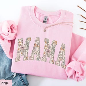 May include: A light pink sweatshirt with the word "MAMA" in large, floral-patterned letters across the front. The sweatshirt is folded, with the sleeves and neckline visible. A necklace with a small pendant rests on the fabric.