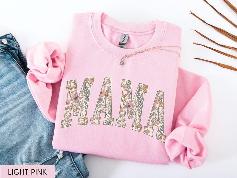 May include: A light pink sweatshirt with the word "MAMA" in large, floral-patterned letters across the front. The sweatshirt is folded, with the sleeves and neckline visible. A necklace with a small pendant rests on the fabric.