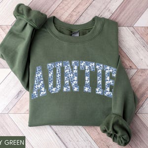 Peut inclure: Sweat-shirt vert militaire avec le mot "AUNTIE" en lettres majuscules blanches remplies d'un motif floral bleu. Le sweat-shirt a un col rond et des manches longues avec des poignets c&ocirc;tel&eacute;s. Le v&ecirc;tement est en mati&egrave;re douce.