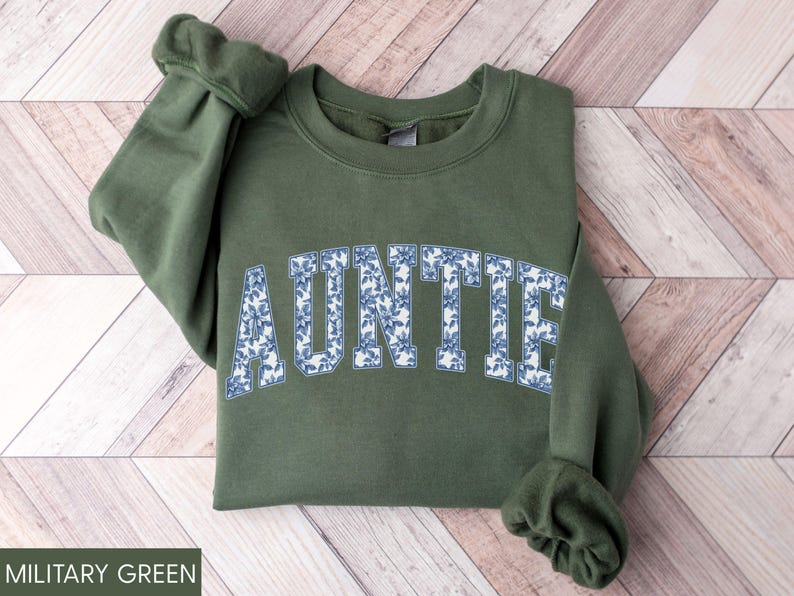 Peut inclure: Sweat-shirt vert militaire avec le mot "AUNTIE" en lettres majuscules blanches remplies d'un motif floral bleu. Le sweat-shirt a un col rond et des manches longues avec des poignets c&ocirc;tel&eacute;s. Le v&ecirc;tement est en mati&egrave;re douce.
