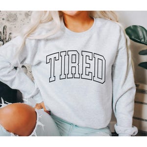 Pu&ograve; includere: Una felpa grigio chiaro con la scritta "TIRED" stampata a grandi lettere nere. La felpa ha un girocollo e maniche lunghe. L'immagine mostra una persona che indossa la felpa.