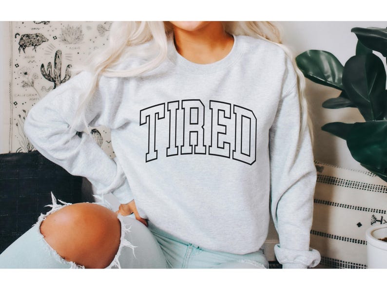 Pu&ograve; includere: Una felpa grigio chiaro con la scritta "TIRED" stampata a grandi lettere nere. La felpa ha un girocollo e maniche lunghe. L'immagine mostra una persona che indossa la felpa.