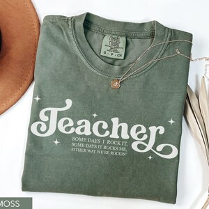 May include: Moss green t-shirt with the word "Teacher" in white script, accented with stars. The shirt also features the text "SOME DAYS I ROCK IT, SOME DAYS IT ROCKS ME, EITHER WAY WE'RE ROCKIN". A gold necklace and a brown hat are also visible.