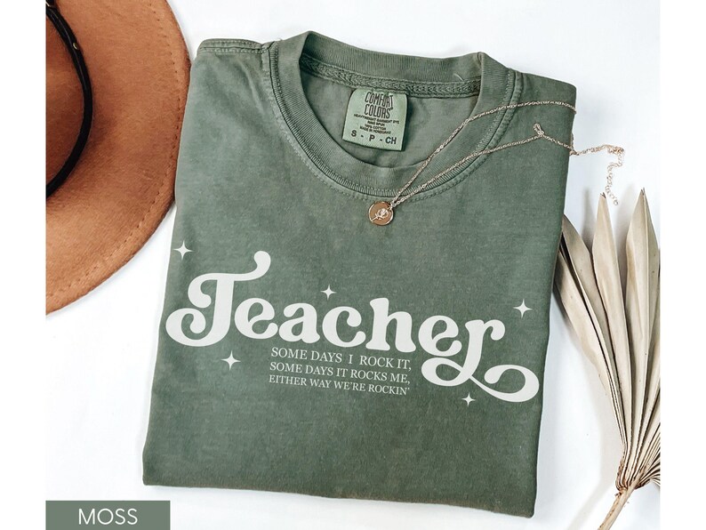 May include: Moss green t-shirt with the word "Teacher" in white script, accented with stars. The shirt also features the text "SOME DAYS I ROCK IT, SOME DAYS IT ROCKS ME, EITHER WAY WE'RE ROCKIN". A gold necklace and a brown hat are also visible.
