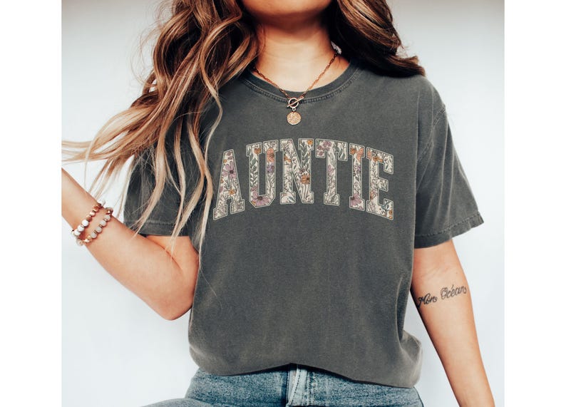 May include: A gray t-shirt with a floral print of the word "AUNTIE" in white letters.
