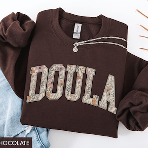 May include: A dark chocolate-colored sweatshirt with the word "DOULA" in a floral design. A silver necklace with a pendant is draped across the sweatshirt. The sweatshirt is folded next to a pair of blue jeans.