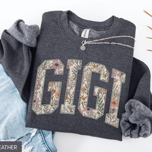 May include: Dark grey sweatshirt featuring the word "GIGI" in a floral design. The letters are filled with flowers in pink, orange, and green. A silver necklace is draped across the sweatshirt. Blue denim jeans are visible.