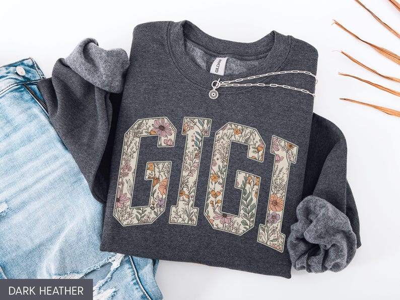 May include: Dark grey sweatshirt featuring the word "GIGI" in a floral design. The letters are filled with flowers in pink, orange, and green. A silver necklace is draped across the sweatshirt. Blue denim jeans are visible.