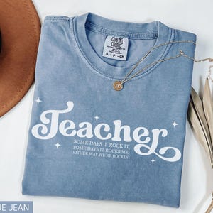 May include: A blue jean Comfort Colors t-shirt with the word "Teacher" in white script and the phrase "SOME DAYS I ROCK IT, SOME DAYS IT ROCKS ME, EITHER WAY WE'RE ROCKIN" in a smaller font. A gold necklace and a brown hat are also visible.