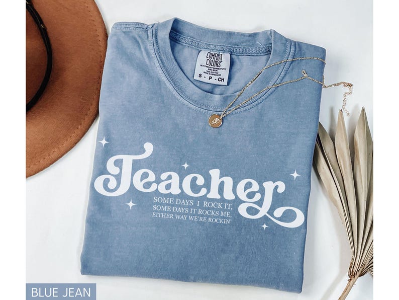 May include: A blue jean Comfort Colors t-shirt with the word "Teacher" in white script and the phrase "SOME DAYS I ROCK IT, SOME DAYS IT ROCKS ME, EITHER WAY WE'RE ROCKIN" in a smaller font. A gold necklace and a brown hat are also visible.