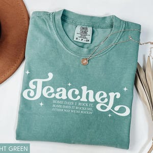 May include: Light green Comfort Colors t-shirt with the word "Teacher" in white script. The shirt also has the text "SOME DAYS I ROCK IT, SOME DAYS IT ROCKS ME, EITHER WAY WE'RE ROCKIN". A gold necklace and a brown hat are also in the image.