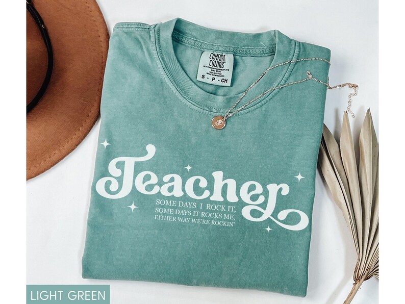 May include: Light green Comfort Colors t-shirt with the word "Teacher" in white script. The shirt also has the text "SOME DAYS I ROCK IT, SOME DAYS IT ROCKS ME, EITHER WAY WE'RE ROCKIN". A gold necklace and a brown hat are also in the image.