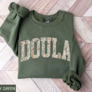May include: Military green sweatshirt with the word "DOULA" in a floral design across the chest. The letters are filled with a pattern of flowers in shades of pink, orange, and cream. The sweatshirt has a crew neck and long sleeves.