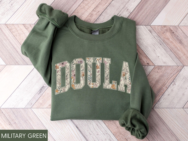 May include: Military green sweatshirt with the word "DOULA" in a floral design across the chest. The letters are filled with a pattern of flowers in shades of pink, orange, and cream. The sweatshirt has a crew neck and long sleeves.