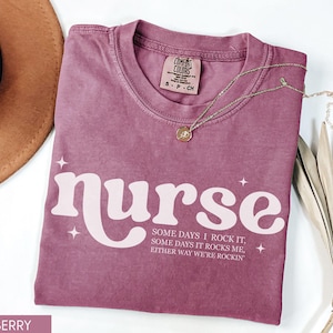 Pode incluir: Camiseta cor de berry com a palavra "nurse" em uma fonte retr&ocirc;, acompanhada do texto "Some days I rock it, some days it rocks me, either way we're rockin". Um colar de ouro e um chap&eacute;u marrom tamb&eacute;m s&atilde;o vis&iacute;veis.
