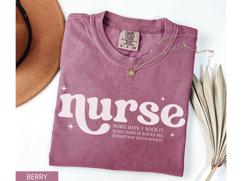 Pode incluir: Camiseta cor de berry com a palavra "nurse" em uma fonte retr&ocirc;, acompanhada do texto "Some days I rock it, some days it rocks me, either way we're rockin". Um colar de ouro e um chap&eacute;u marrom tamb&eacute;m s&atilde;o vis&iacute;veis.