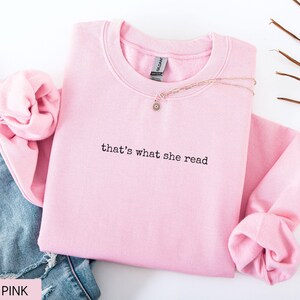 May include: A light pink sweatshirt with the phrase "that's what she read" in black lettering. A gold necklace with a small pendant rests on the sweatshirt. The sweatshirt is folded next to a pair of blue jeans.