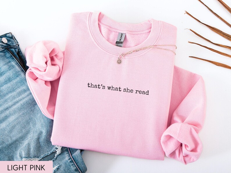 May include: A light pink sweatshirt with the phrase "that's what she read" in black lettering. A gold necklace with a small pendant rests on the sweatshirt. The sweatshirt is folded next to a pair of blue jeans.