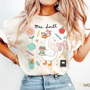 May include: Ivory-colored t-shirt featuring a cartoon goose wearing glasses and a bow, carrying books. The design includes pencils, an apple, and the text "Mrs. Smith". A cute and colorful teacher-themed graphic.